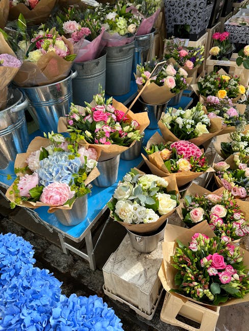 A display of freshly arranged flower bouquets, featuring pink, white, and blue blooms such as roses, hydrangeas, and carnations, wrapped in brown craft paper and set in metal buckets and vases. The bo