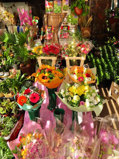 A vibrant display of fresh floral bouquets arranged on a pink table in a flower shop. The bouquets feature a variety of colorful flowers, including bright yellow, pink, red, and white blooms, wrapped 