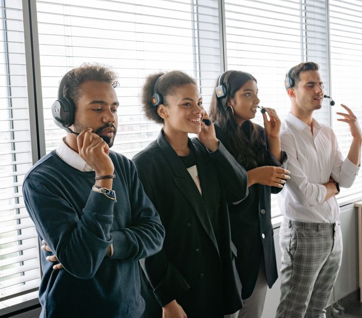 A diverse group of four customer service representatives wearing headsets, standing in front of large window blinds in an office environment. The first person, a man with curly hair and a dark blue sw