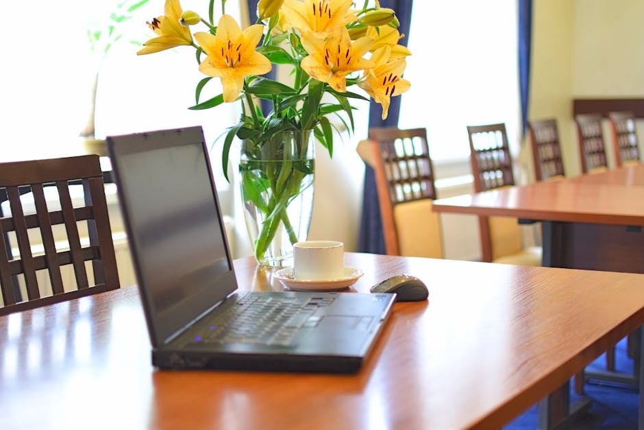 A glass vase filled with vibrant yellow lilies with elongated petals and dark stamens, arranged in a lush bouquet on a wooden office table. The bouquet is surrounded by green foliage, with a white tea