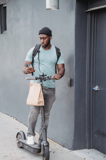 A young man with dark skin, wearing a black beanie, light blue t-shirt, and grey jeans, is standing outside a grey building wall next to a black door. He is holding a smartphone in his right hand and 