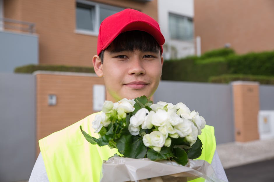 A young delivery person wearing a red cap and a high-visibility yellow vest is outdoors, holding a bouquet of white flowers with green leaves wrapped in white paper. The flowers appear fresh with soft